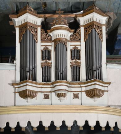 Zdjęcie nr 1: Chór muzyczny stanowi przyścienną, trójosiową, konstrukcję zawieszoną na dwóch kroksztynach i wspartą na dwóch smukłych kolumnach podtrzymujących oś środkową. Prowadzą na niego dwubiegowe schody w nawie ustawione od południa. Parapet chóru jest trójosiowy, wybrzuszony w osi środkowej, z silnie zaznaczonym, wyłamującym się cokołem i podwieszonym lambrekinem. W osiach bocznych parapet dekorują trzy malowane okręgi z przestylizowanym krzyżem wewnątrz, otoczonym potrójnymi promieniami pomiędzy ramionami, poprzedzielane smukłymi gałązkami kwiatów, z wyobrażeniem trawy poniżej. Prospekt jest pięcioosiowy, z osiami skrajnymi wyższymi od środkowej, podtrzymywany przez trzy konsolki z imitacją puklowania. Cokół wsparty na konsolach jest wyłamany na osiach skrajnych i wypukły w osi środkowej. Nisze na piszczałki są prostokątne. Siedem piszczałek ustawiono pod kątem. Zbiegają się  one ku środkowi. W górnej partii umieszczono ornament utworzony ze stylizowanej wici roślinnej. Powyżej znajduje się niepełne belkowanie, wyłamane analogicznie jak cokół, z pionowymi prześwitami, zwieńczone ornamentami roślinnymi i krzyżem pośrodku. Osie druga i czwarta są dwukondygnacyjne z prostokątnymi prześwitami na piszczałki, ze zwieńczeniami utworzonymi z ze stylizowanej wici roślinnej. Kolorystyka prospektu jest jasna, beżowa. Detale, cokoły i belkowania pierwotnie były złocone.
