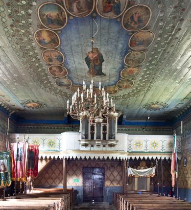 Zdjęcie nr 2: Chór muzyczny stanowi przyścienną, trójosiową, konstrukcję zawieszoną na dwóch kroksztynach i wspartą na dwóch smukłych kolumnach podtrzymujących oś środkową. Prowadzą na niego dwubiegowe schody w nawie ustawione od południa. Parapet chóru jest trójosiowy, wybrzuszony w osi środkowej, z silnie zaznaczonym, wyłamującym się cokołem i podwieszonym lambrekinem. W osiach bocznych parapet dekorują trzy malowane okręgi z przestylizowanym krzyżem wewnątrz, otoczonym potrójnymi promieniami pomiędzy ramionami, poprzedzielane smukłymi gałązkami kwiatów, z wyobrażeniem trawy poniżej. Prospekt jest pięcioosiowy, z osiami skrajnymi wyższymi od środkowej, podtrzymywany przez trzy konsolki z imitacją puklowania. Cokół wsparty na konsolach jest wyłamany na osiach skrajnych i wypukły w osi środkowej. Nisze na piszczałki są prostokątne. Siedem piszczałek ustawiono pod kątem. Zbiegają się  one ku środkowi. W górnej partii umieszczono ornament utworzony ze stylizowanej wici roślinnej. Powyżej znajduje się niepełne belkowanie, wyłamane analogicznie jak cokół, z pionowymi prześwitami, zwieńczone ornamentami roślinnymi i krzyżem pośrodku. Osie druga i czwarta są dwukondygnacyjne z prostokątnymi prześwitami na piszczałki, ze zwieńczeniami utworzonymi z ze stylizowanej wici roślinnej. Kolorystyka prospektu jest jasna, beżowa. Detale, cokoły i belkowania pierwotnie były złocone.
