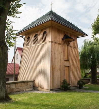 Zdjęcie nr 2: Dzwonnica drewniana, konstrukcji słupowej, ściany szalowane z listowaniem, więźba dachowa drewniana. Założona na planie prostokąta, na kamiennym podmurowaniu. Ściany zwężające się ku górze, odcięte listwami w trzech czwartych wysokości. Dach gontowy, czterospadowy, zakończony krzyżem. Otwór drzwiowy w ścianie południowej o wykroju stojącego prostokąta zamknięty drzwiami szalowanymi z listowaniem. Ponad nimi, na osi otwór w formie stojącego prostokąta zamkniętego łukiem odcinkowym, zamknięty drzwiczkami szalowanymi z listowaniem. Otwór nakryty gontowym daszkiem okapowym. W najwyższej partii ścian otwory głosowe w formie stojącego prostokąta zamkniętego łukiem półkolistym nadwieszonym po trzy na wschodniej i zachodniej ścianie i jednym na północnej. 