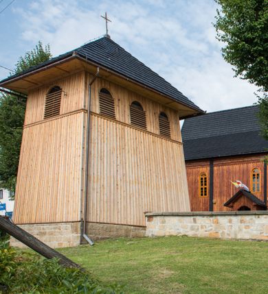 Zdjęcie nr 1: Dzwonnica drewniana, konstrukcji słupowej, ściany szalowane z listowaniem, więźba dachowa drewniana. Założona na planie prostokąta, na kamiennym podmurowaniu. Ściany zwężające się ku górze, odcięte listwami w trzech czwartych wysokości. Dach gontowy, czterospadowy, zakończony krzyżem. Otwór drzwiowy w ścianie południowej o wykroju stojącego prostokąta zamknięty drzwiami szalowanymi z listowaniem. Ponad nimi, na osi otwór w formie stojącego prostokąta zamkniętego łukiem odcinkowym, zamknięty drzwiczkami szalowanymi z listowaniem. Otwór nakryty gontowym daszkiem okapowym. W najwyższej partii ścian otwory głosowe w formie stojącego prostokąta zamkniętego łukiem półkolistym nadwieszonym po trzy na wschodniej i zachodniej ścianie i jednym na północnej. 