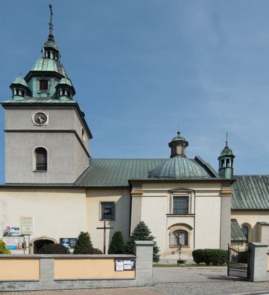 Zdjęcie nr 1: Kościół orientowany, jest budowlą murowaną z cegły i kamienia, na rzucie podłużnym. Korpus trójnawowy i dwuprzęsłowy. Prezbiterium trójprzęsłowe, zamknięte trójbocznie prezbiterium z przylegającymi od północy zakrystią, od południa kruchtą. Na osi kościoła, jednokondygnacyjna, szeroka, przysadzista wieża z kruchtą w przyziemiu i przyległością od zachodu.  Od południa i zachodu, w obrębie pierwszego przęsła naw bocznych jest para kaplic na rzutach zbliżonych do kwadratu. Nawa i prezbiterium przekryte są sklepieniami kolebkowymi z lunetami wspartymi na pilastrach i filarach. Kaplice wieńczą kopuły. Artykulację ścian na zewnątrz prezbiterium stanowi zespół przypór, naroża kaplic ujmują pilastry wpierające obiegający bryłę niepełne belkowanie.
Do wnętrza prowadzi 6 otworów drzwiowych, z czego 2 w kruchcie w wieży ujęte kamiennymi portalami zamkniętymi łukiem koszowym i do kaplicy ujętym kamiennym portalem w formie pary lizen na wysokich cokołach wspierających belkowanie z parą rozet w partii fryzu. Okna korpusu nawowego w kształcie stojących prostokątów, prezbiterium ostrołukowe, natomiast kaplic są prostokątne ujęte kamiennym obramieniem z uszakami w górnych narożach i zwieńczone trójkątnymi przyczółkami. Wieża artykułowana w 2/3 wysokości gzymsem. W dolnej strefie w 3 z 4 ścian po jednym, zamkniętym półkoliście oknie. W górnej partii pośrodku  z czterech po 1 okrągłej tarczy zegara. Wieże wieńczy hełm ujęty 4 latarniami i zwieńczony piątą. Dachy korpusu i prezbiterium dwuspadowe, nad kaplicami kopuły z latarniami, nad zakrystią i kruchtą boczną pulpitowe. Tynki ścian w odcieniach bieli i szarości. 
