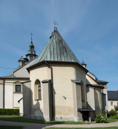 Zdjęcie nr 18: Kościół orientowany, jest budowlą murowaną z cegły i kamienia, na rzucie podłużnym. Korpus trójnawowy i dwuprzęsłowy. Prezbiterium trójprzęsłowe, zamknięte trójbocznie prezbiterium z przylegającymi od północy zakrystią, od południa kruchtą. Na osi kościoła, jednokondygnacyjna, szeroka, przysadzista wieża z kruchtą w przyziemiu i przyległością od zachodu.  Od południa i zachodu, w obrębie pierwszego przęsła naw bocznych jest para kaplic na rzutach zbliżonych do kwadratu. Nawa i prezbiterium przekryte są sklepieniami kolebkowymi z lunetami wspartymi na pilastrach i filarach. Kaplice wieńczą kopuły. Artykulację ścian na zewnątrz prezbiterium stanowi zespół przypór, naroża kaplic ujmują pilastry wpierające obiegający bryłę niepełne belkowanie.
Do wnętrza prowadzi 6 otworów drzwiowych, z czego 2 w kruchcie w wieży ujęte kamiennymi portalami zamkniętymi łukiem koszowym i do kaplicy ujętym kamiennym portalem w formie pary lizen na wysokich cokołach wspierających belkowanie z parą rozet w partii fryzu. Okna korpusu nawowego w kształcie stojących prostokątów, prezbiterium ostrołukowe, natomiast kaplic są prostokątne ujęte kamiennym obramieniem z uszakami w górnych narożach i zwieńczone trójkątnymi przyczółkami. Wieża artykułowana w 2/3 wysokości gzymsem. W dolnej strefie w 3 z 4 ścian po jednym, zamkniętym półkoliście oknie. W górnej partii pośrodku  z czterech po 1 okrągłej tarczy zegara. Wieże wieńczy hełm ujęty 4 latarniami i zwieńczony piątą. Dachy korpusu i prezbiterium dwuspadowe, nad kaplicami kopuły z latarniami, nad zakrystią i kruchtą boczną pulpitowe. Tynki ścian w odcieniach bieli i szarości. 

