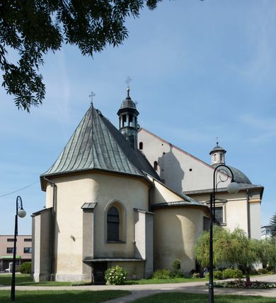 Zdjęcie nr 13: Kościół orientowany, jest budowlą murowaną z cegły i kamienia, na rzucie podłużnym. Korpus trójnawowy i dwuprzęsłowy. Prezbiterium trójprzęsłowe, zamknięte trójbocznie prezbiterium z przylegającymi od północy zakrystią, od południa kruchtą. Na osi kościoła, jednokondygnacyjna, szeroka, przysadzista wieża z kruchtą w przyziemiu i przyległością od zachodu.  Od południa i zachodu, w obrębie pierwszego przęsła naw bocznych jest para kaplic na rzutach zbliżonych do kwadratu. Nawa i prezbiterium przekryte są sklepieniami kolebkowymi z lunetami wspartymi na pilastrach i filarach. Kaplice wieńczą kopuły. Artykulację ścian na zewnątrz prezbiterium stanowi zespół przypór, naroża kaplic ujmują pilastry wpierające obiegający bryłę niepełne belkowanie.
Do wnętrza prowadzi 6 otworów drzwiowych, z czego 2 w kruchcie w wieży ujęte kamiennymi portalami zamkniętymi łukiem koszowym i do kaplicy ujętym kamiennym portalem w formie pary lizen na wysokich cokołach wspierających belkowanie z parą rozet w partii fryzu. Okna korpusu nawowego w kształcie stojących prostokątów, prezbiterium ostrołukowe, natomiast kaplic są prostokątne ujęte kamiennym obramieniem z uszakami w górnych narożach i zwieńczone trójkątnymi przyczółkami. Wieża artykułowana w 2/3 wysokości gzymsem. W dolnej strefie w 3 z 4 ścian po jednym, zamkniętym półkoliście oknie. W górnej partii pośrodku  z czterech po 1 okrągłej tarczy zegara. Wieże wieńczy hełm ujęty 4 latarniami i zwieńczony piątą. Dachy korpusu i prezbiterium dwuspadowe, nad kaplicami kopuły z latarniami, nad zakrystią i kruchtą boczną pulpitowe. Tynki ścian w odcieniach bieli i szarości. 

