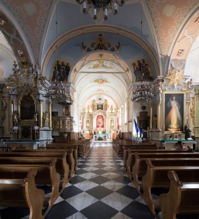 Zdjęcie nr 9: Kościół orientowany, jest budowlą murowaną z cegły i kamienia, na rzucie podłużnym. Korpus trójnawowy i dwuprzęsłowy. Prezbiterium trójprzęsłowe, zamknięte trójbocznie prezbiterium z przylegającymi od północy zakrystią, od południa kruchtą. Na osi kościoła, jednokondygnacyjna, szeroka, przysadzista wieża z kruchtą w przyziemiu i przyległością od zachodu.  Od południa i zachodu, w obrębie pierwszego przęsła naw bocznych jest para kaplic na rzutach zbliżonych do kwadratu. Nawa i prezbiterium przekryte są sklepieniami kolebkowymi z lunetami wspartymi na pilastrach i filarach. Kaplice wieńczą kopuły. Artykulację ścian na zewnątrz prezbiterium stanowi zespół przypór, naroża kaplic ujmują pilastry wpierające obiegający bryłę niepełne belkowanie.
Do wnętrza prowadzi 6 otworów drzwiowych, z czego 2 w kruchcie w wieży ujęte kamiennymi portalami zamkniętymi łukiem koszowym i do kaplicy ujętym kamiennym portalem w formie pary lizen na wysokich cokołach wspierających belkowanie z parą rozet w partii fryzu. Okna korpusu nawowego w kształcie stojących prostokątów, prezbiterium ostrołukowe, natomiast kaplic są prostokątne ujęte kamiennym obramieniem z uszakami w górnych narożach i zwieńczone trójkątnymi przyczółkami. Wieża artykułowana w 2/3 wysokości gzymsem. W dolnej strefie w 3 z 4 ścian po jednym, zamkniętym półkoliście oknie. W górnej partii pośrodku  z czterech po 1 okrągłej tarczy zegara. Wieże wieńczy hełm ujęty 4 latarniami i zwieńczony piątą. Dachy korpusu i prezbiterium dwuspadowe, nad kaplicami kopuły z latarniami, nad zakrystią i kruchtą boczną pulpitowe. Tynki ścian w odcieniach bieli i szarości. 
