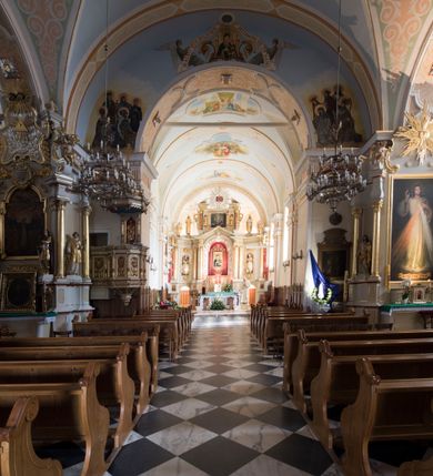 Zdjęcie nr 8: Kościół orientowany, jest budowlą murowaną z cegły i kamienia, na rzucie podłużnym. Korpus trójnawowy i dwuprzęsłowy. Prezbiterium trójprzęsłowe, zamknięte trójbocznie prezbiterium z przylegającymi od północy zakrystią, od południa kruchtą. Na osi kościoła, jednokondygnacyjna, szeroka, przysadzista wieża z kruchtą w przyziemiu i przyległością od zachodu.  Od południa i zachodu, w obrębie pierwszego przęsła naw bocznych jest para kaplic na rzutach zbliżonych do kwadratu. Nawa i prezbiterium przekryte są sklepieniami kolebkowymi z lunetami wspartymi na pilastrach i filarach. Kaplice wieńczą kopuły. Artykulację ścian na zewnątrz prezbiterium stanowi zespół przypór, naroża kaplic ujmują pilastry wpierające obiegający bryłę niepełne belkowanie.
Do wnętrza prowadzi 6 otworów drzwiowych, z czego 2 w kruchcie w wieży ujęte kamiennymi portalami zamkniętymi łukiem koszowym i do kaplicy ujętym kamiennym portalem w formie pary lizen na wysokich cokołach wspierających belkowanie z parą rozet w partii fryzu. Okna korpusu nawowego w kształcie stojących prostokątów, prezbiterium ostrołukowe, natomiast kaplic są prostokątne ujęte kamiennym obramieniem z uszakami w górnych narożach i zwieńczone trójkątnymi przyczółkami. Wieża artykułowana w 2/3 wysokości gzymsem. W dolnej strefie w 3 z 4 ścian po jednym, zamkniętym półkoliście oknie. W górnej partii pośrodku  z czterech po 1 okrągłej tarczy zegara. Wieże wieńczy hełm ujęty 4 latarniami i zwieńczony piątą. Dachy korpusu i prezbiterium dwuspadowe, nad kaplicami kopuły z latarniami, nad zakrystią i kruchtą boczną pulpitowe. Tynki ścian w odcieniach bieli i szarości. 

