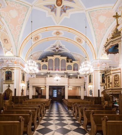 Zdjęcie nr 15: Kościół orientowany, jest budowlą murowaną z cegły i kamienia, na rzucie podłużnym. Korpus trójnawowy i dwuprzęsłowy. Prezbiterium trójprzęsłowe, zamknięte trójbocznie prezbiterium z przylegającymi od północy zakrystią, od południa kruchtą. Na osi kościoła, jednokondygnacyjna, szeroka, przysadzista wieża z kruchtą w przyziemiu i przyległością od zachodu.  Od południa i zachodu, w obrębie pierwszego przęsła naw bocznych jest para kaplic na rzutach zbliżonych do kwadratu. Nawa i prezbiterium przekryte są sklepieniami kolebkowymi z lunetami wspartymi na pilastrach i filarach. Kaplice wieńczą kopuły. Artykulację ścian na zewnątrz prezbiterium stanowi zespół przypór, naroża kaplic ujmują pilastry wpierające obiegający bryłę niepełne belkowanie.
Do wnętrza prowadzi 6 otworów drzwiowych, z czego 2 w kruchcie w wieży ujęte kamiennymi portalami zamkniętymi łukiem koszowym i do kaplicy ujętym kamiennym portalem w formie pary lizen na wysokich cokołach wspierających belkowanie z parą rozet w partii fryzu. Okna korpusu nawowego w kształcie stojących prostokątów, prezbiterium ostrołukowe, natomiast kaplic są prostokątne ujęte kamiennym obramieniem z uszakami w górnych narożach i zwieńczone trójkątnymi przyczółkami. Wieża artykułowana w 2/3 wysokości gzymsem. W dolnej strefie w 3 z 4 ścian po jednym, zamkniętym półkoliście oknie. W górnej partii pośrodku  z czterech po 1 okrągłej tarczy zegara. Wieże wieńczy hełm ujęty 4 latarniami i zwieńczony piątą. Dachy korpusu i prezbiterium dwuspadowe, nad kaplicami kopuły z latarniami, nad zakrystią i kruchtą boczną pulpitowe. Tynki ścian w odcieniach bieli i szarości. 
