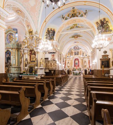 Zdjęcie nr 12: Kościół orientowany, jest budowlą murowaną z cegły i kamienia, na rzucie podłużnym. Korpus trójnawowy i dwuprzęsłowy. Prezbiterium trójprzęsłowe, zamknięte trójbocznie prezbiterium z przylegającymi od północy zakrystią, od południa kruchtą. Na osi kościoła, jednokondygnacyjna, szeroka, przysadzista wieża z kruchtą w przyziemiu i przyległością od zachodu.  Od południa i zachodu, w obrębie pierwszego przęsła naw bocznych jest para kaplic na rzutach zbliżonych do kwadratu. Nawa i prezbiterium przekryte są sklepieniami kolebkowymi z lunetami wspartymi na pilastrach i filarach. Kaplice wieńczą kopuły. Artykulację ścian na zewnątrz prezbiterium stanowi zespół przypór, naroża kaplic ujmują pilastry wpierające obiegający bryłę niepełne belkowanie.
Do wnętrza prowadzi 6 otworów drzwiowych, z czego 2 w kruchcie w wieży ujęte kamiennymi portalami zamkniętymi łukiem koszowym i do kaplicy ujętym kamiennym portalem w formie pary lizen na wysokich cokołach wspierających belkowanie z parą rozet w partii fryzu. Okna korpusu nawowego w kształcie stojących prostokątów, prezbiterium ostrołukowe, natomiast kaplic są prostokątne ujęte kamiennym obramieniem z uszakami w górnych narożach i zwieńczone trójkątnymi przyczółkami. Wieża artykułowana w 2/3 wysokości gzymsem. W dolnej strefie w 3 z 4 ścian po jednym, zamkniętym półkoliście oknie. W górnej partii pośrodku  z czterech po 1 okrągłej tarczy zegara. Wieże wieńczy hełm ujęty 4 latarniami i zwieńczony piątą. Dachy korpusu i prezbiterium dwuspadowe, nad kaplicami kopuły z latarniami, nad zakrystią i kruchtą boczną pulpitowe. Tynki ścian w odcieniach bieli i szarości. 
