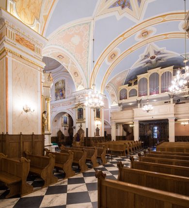 Zdjęcie nr 17: Kościół orientowany, jest budowlą murowaną z cegły i kamienia, na rzucie podłużnym. Korpus trójnawowy i dwuprzęsłowy. Prezbiterium trójprzęsłowe, zamknięte trójbocznie prezbiterium z przylegającymi od północy zakrystią, od południa kruchtą. Na osi kościoła, jednokondygnacyjna, szeroka, przysadzista wieża z kruchtą w przyziemiu i przyległością od zachodu.  Od południa i zachodu, w obrębie pierwszego przęsła naw bocznych jest para kaplic na rzutach zbliżonych do kwadratu. Nawa i prezbiterium przekryte są sklepieniami kolebkowymi z lunetami wspartymi na pilastrach i filarach. Kaplice wieńczą kopuły. Artykulację ścian na zewnątrz prezbiterium stanowi zespół przypór, naroża kaplic ujmują pilastry wpierające obiegający bryłę niepełne belkowanie.
Do wnętrza prowadzi 6 otworów drzwiowych, z czego 2 w kruchcie w wieży ujęte kamiennymi portalami zamkniętymi łukiem koszowym i do kaplicy ujętym kamiennym portalem w formie pary lizen na wysokich cokołach wspierających belkowanie z parą rozet w partii fryzu. Okna korpusu nawowego w kształcie stojących prostokątów, prezbiterium ostrołukowe, natomiast kaplic są prostokątne ujęte kamiennym obramieniem z uszakami w górnych narożach i zwieńczone trójkątnymi przyczółkami. Wieża artykułowana w 2/3 wysokości gzymsem. W dolnej strefie w 3 z 4 ścian po jednym, zamkniętym półkoliście oknie. W górnej partii pośrodku  z czterech po 1 okrągłej tarczy zegara. Wieże wieńczy hełm ujęty 4 latarniami i zwieńczony piątą. Dachy korpusu i prezbiterium dwuspadowe, nad kaplicami kopuły z latarniami, nad zakrystią i kruchtą boczną pulpitowe. Tynki ścian w odcieniach bieli i szarości. 
