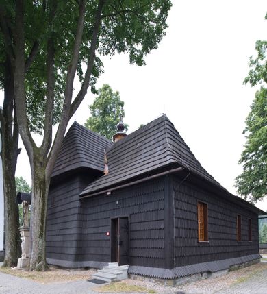 Zdjęcie nr 13: Kościół drewniany, orientowany, o konstrukcji zrębowej, obity gontami. Budowla jednonawowa, z węższym prezbiterium zamkniętym prosto, z zakrystią i kaplicą od strony północnej i kruchtą od południa. Nawa prostokątna, bardzo wydłużona, prezbiterium krótkie, zamknięte prosto. Wszystkie ściany i strony pokrywa polichromia o motywach ornamentalnych i roślinnych otaczających sceny figuralne. Nawa przekryta stropem z zaskrzynieniami, co oznacza, że część konstrukcji dachu, która znajduje się już w przestrzeni nawy została zakryta stropem o skośnym przebiegu, a prezbiterium pozornym stropem kolebkowym. Po prawej stronie dwa duże okna w nawie i jedno w prezbiterium, trzy kolejne w kaplicy. Wejście główne od zachodu, kolejne przez południową kruchtę i do kaplicy, w ścianie zakrystii barokowy portal kamienny z ornamentem roślinnym. Ściany zewnętrzne gładkie, posadowione na niewysokim podmurowaniu zakrytym gontowym fartuchem. Dachy gontowe o konstrukcji więźbowo-zaskrzyniowej. Na kalenicy wieżyczka na sygnaturkę z baniastym hełmem krytym blachą. 