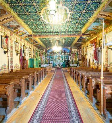 Zdjęcie nr 19: Kościół drewniany, orientowany, o konstrukcji zrębowej, obity gontami. Budowla jednonawowa, z węższym prezbiterium zamkniętym prosto, z zakrystią i kaplicą od strony północnej i kruchtą od południa. Nawa prostokątna, bardzo wydłużona, prezbiterium krótkie, zamknięte prosto. Wszystkie ściany i strony pokrywa polichromia o motywach ornamentalnych i roślinnych otaczających sceny figuralne. Nawa przekryta stropem z zaskrzynieniami, co oznacza, że część konstrukcji dachu, która znajduje się już w przestrzeni nawy została zakryta stropem o skośnym przebiegu, a prezbiterium pozornym stropem kolebkowym. Po prawej stronie dwa duże okna w nawie i jedno w prezbiterium, trzy kolejne w kaplicy. Wejście główne od zachodu, kolejne przez południową kruchtę i do kaplicy, w ścianie zakrystii barokowy portal kamienny z ornamentem roślinnym. Ściany zewnętrzne gładkie, posadowione na niewysokim podmurowaniu zakrytym gontowym fartuchem. Dachy gontowe o konstrukcji więźbowo-zaskrzyniowej. Na kalenicy wieżyczka na sygnaturkę z baniastym hełmem krytym blachą. 