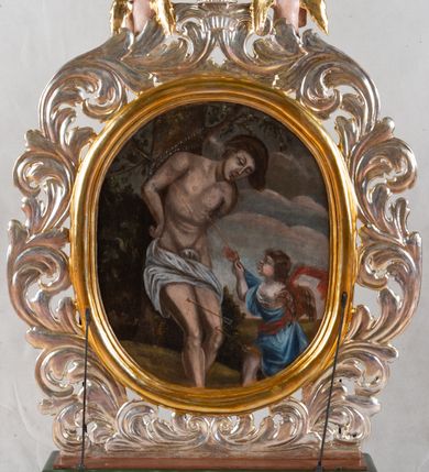 Zdjęcie nr 3: Feretron dwustronny z przedstawieniem Matki Bożej i św. Sebastiana. Na profilowanym postumencie zaopatrzonym w otwory na drążki do noszenia, dołem puklowanym, została umieszczona dwustronnie rzeźbiona, owalna rama utworzona z liści schnącego akantu, z dwoma aniołkami podtrzymującymi koronę w zwieńczeniu. Aniołki ukazano frontalnie, z jedną ręką złożoną na brzuchu, drugą wyprostowaną, podtrzymującą koronę w typie zamkniętym, ze złożonymi skrzydłami, z przepaską na biodrach utworzoną z liści akantu. Mają jasną karnację, szerokie twarze, zarumienione policzki, drobne, czerwone usta oraz krótkie, szerokie nosy. Włosy ciemnobrązowe, średniej długości, z przedziałkiem na środku. Na awersie znajduje się obraz z wizerunkiem popiersia Matki Bożej w owalnej ramie utworzonej z liści przewiązanych w czterech miejscach karbowaną wstęgą. Maria ukazana frontalnie, z głową delikatnie przechyloną w dół i zwróconą w prawo, z rękami złożonymi na piersi. Ubrana jest w białą suknię z długimi rękawami i niebieski płaszcz zarzucony na ramiona. Na głowie ma białą chustę i niebieski welon. Włosy długie, z przedziałkiem na środku, falowane. Dłonie subtelne, z długimi palcami, karnacja jasna, drobne usta, przymknięte powieki, długi, wąski nos. Wokół głowy Marii namalowano 11 gwiazd. Tło obrazu jasnożółte. Na rewersie obraz z przedstawieniem św. Sebastiana w owalnej, profilowanej, wypukłej ramie. W centrum kompozycji postać męczennika przywiązanego do drzewa. Jest zwrócony w trzech czwartych w lewo, delikatnie pochylony, z nogami ugiętymi w kolanach, rękami związanymi z tyłu. Ciało przebite czterema strzałami tkwiącymi w prawym ramieniu, lewym boku i obydwu nogach. Twarz podłużna z długim, wąskim nosem. U dołu po prawej aniołek, wysokością sięgający do połowy św. Sebastiana, ukazany z lewego profilu, wyciągający strzałę z boku świętego. Aniołek jest ubrany w niebieską szatę do kolan oraz czerwoną chustę zwiewnie unoszącą się ponad plecami. Ma twarz okrągłą, z wysuniętym podbródkiem, duże, szerokie usta, krótki, wąski nos, włosy średniej długości. W tle, po lewej stronie roślinność, po prawej błękitne niebo. Polichromia podstawy w kolorze brązowym i zielonym, rama i detale złocone i srebrzone.