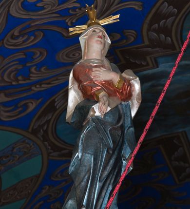 Zdjęcie nr 3: Rzeźby ukrzyżowanego Jezusa, Matki Boskiej oraz św. Jana Ewangelisty tworzące grupę Ukrzyżowania. W centrum kompozycji krzyż o trójlistnym zakończeniu ramion, na nim figura Chrystusa w typie Christo morte, przybita do krzyża trzema gwoździami. Ciało w delikatnym zwisie o stosunkowo szeroko rozłożonych ramionach, esowato wygięte, kolana zgięte, stopy w układzie prawa na lewą, głowa opada na prawy bark. Jezus ma szczupłą twarz, okoloną krótkim zarostem oraz długimi, silnie skręconymi włosami opadającymi na plecy i prawe ramię; na głowie gruba, zielona korona cierniowa. Ciało silnie wychudzone o wyraźnie podkreślonej muskulaturze oraz zapadniętej klatce piersiowej z zaznaczoną linią żeber oraz mostka. Przez biodra przewiązane krótkim, złotym perizonium zawiązanym w partii podbrzusza, z fragmentami tkaniny rozwianymi na boki, draperia silnie łamana. 
Po lewej stronie krzyża figura Matki Boskiej, zwrócona w stronę Ukrzyżowanego. Maria stoi w kontrapoście, ma esowato wygięto sylwetkę i głowę uniesioną do góry. Jej lewa ręka spoczywa na piersi, prawą wyciąga przed siebie. Ma pełną twarz, o wystającym podbródku. Ubrana jest w czerwoną suknię o wywiniętych mankietach oraz okalający jej ciało niebieski płaszcz; na głowie ma biały welon. 
Po prawej stronie figura św. Jana ukazanego w skręcie, z rękami złożonymi  po lewej stronie ciała i z głową skierowaną w prawo. Święty stoi w kontrapoście, z prawym biodrem mocno wypchniętym do boku. Ma prostokątną twarz z półprzymkniętymi, migdałowatymi oczami, dużym nosem i mocno zarysowanymi ustami, wzrok kieruje w dół. Na plecy i ramiona opadają mu silnie skręcone, długie włosy. Jest ubrany w laserowaną zieloną suknię z żółtym podbiciem, silnie przylegającą do ciała i marszczoną oraz czerwony płaszcz przerzucony przez lewe ramię, opadający na lewy bok i plecy. Polichromia naturalistyczna w odsłoniętych partiach ciała. 