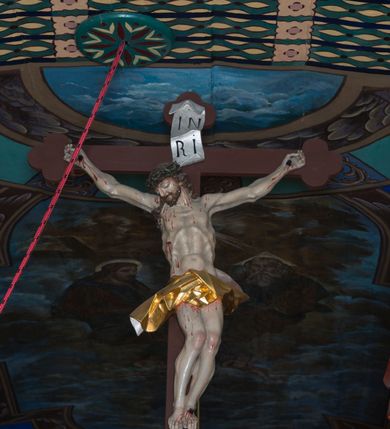 Zdjęcie nr 2: Rzeźby ukrzyżowanego Jezusa, Matki Boskiej oraz św. Jana Ewangelisty tworzące grupę Ukrzyżowania. W centrum kompozycji krzyż o trójlistnym zakończeniu ramion, na nim figura Chrystusa w typie Christo morte, przybita do krzyża trzema gwoździami. Ciało w delikatnym zwisie o stosunkowo szeroko rozłożonych ramionach, esowato wygięte, kolana zgięte, stopy w układzie prawa na lewą, głowa opada na prawy bark. Jezus ma szczupłą twarz, okoloną krótkim zarostem oraz długimi, silnie skręconymi włosami opadającymi na plecy i prawe ramię; na głowie gruba, zielona korona cierniowa. Ciało silnie wychudzone o wyraźnie podkreślonej muskulaturze oraz zapadniętej klatce piersiowej z zaznaczoną linią żeber oraz mostka. Przez biodra przewiązane krótkim, złotym perizonium zawiązanym w partii podbrzusza, z fragmentami tkaniny rozwianymi na boki, draperia silnie łamana. 
Po lewej stronie krzyża figura Matki Boskiej, zwrócona w stronę Ukrzyżowanego. Maria stoi w kontrapoście, ma esowato wygięto sylwetkę i głowę uniesioną do góry. Jej lewa ręka spoczywa na piersi, prawą wyciąga przed siebie. Ma pełną twarz, o wystającym podbródku. Ubrana jest w czerwoną suknię o wywiniętych mankietach oraz okalający jej ciało niebieski płaszcz; na głowie ma biały welon. 
Po prawej stronie figura św. Jana ukazanego w skręcie, z rękami złożonymi  po lewej stronie ciała i z głową skierowaną w prawo. Święty stoi w kontrapoście, z prawym biodrem mocno wypchniętym do boku. Ma prostokątną twarz z półprzymkniętymi, migdałowatymi oczami, dużym nosem i mocno zarysowanymi ustami, wzrok kieruje w dół. Na plecy i ramiona opadają mu silnie skręcone, długie włosy. Jest ubrany w laserowaną zieloną suknię z żółtym podbiciem, silnie przylegającą do ciała i marszczoną oraz czerwony płaszcz przerzucony przez lewe ramię, opadający na lewy bok i plecy. Polichromia naturalistyczna w odsłoniętych partiach ciała. 