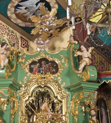 Zdjęcie nr 2: Nastawa ołtarzowa architektoniczna, jednoosiowa, jednokondygnacyjna ze zwieńczeniem. Mensa sarkofagowa, dekorowana na froncie dwiema płycinami w kształcie leżących prostokątów, ujętych po bokach i spiętych motywem ornamentalnym utworzonym z rocaille’a i liści.  
 Oś nastawy flankowana pilastrami przechodzącymi dołem w konsole i górą we fragmenty pełnego belkowania, o delikatnie wklęsło-wypukłej linii gzymsu. Zwieńczenie w formie wklęsło-wypukłej ujęte spływającymi na belkowanie dużymi wolutami, zamknięte stylizowanym gzymsem z ceowników i zwieńczone gołębicą Ducha Świętego na tle glorii, między główkami anielskimi, osadzone na obłoku. W polu środkowym pierwszej kondygnacji nisza z figurą Matki Boskiej Różańcowej wymiennie z obrazem ze sceną Zwiastowania ujęta profilowaną ramą w formie stojącego prostokąta zamkniętego łukiem półkolistym nadwieszonym z uskokiem, dekorowana fragmentami rocaille’ów. Na konsolach figury św. Joachima po lewej stronie i św. Józefa po prawej. W polu zwieńczenia obraz z przedstawieniem św. Rodziny ujęty ramą w formie trójlistnej, dekorowaną fragmentami rocaille'ów i flankowaną kwitnącymi gałązkami. Brzeg zwieńczenia i woluty zdobione taśmą i rocaille’ami, na wolutach para siedzących puttów, na zwieńczeniu uskrzydlone główki anielskie. 
Struktura polichromowana w kolorze zielonym, z marmoryzowanym cokołem i płycinami; ornamentyka i profile złocone i srebrzone.
