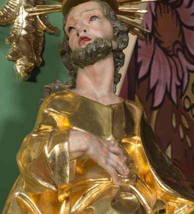 Zdjęcie nr 4: Rzeźba pełna, na cokole, ukazująca św. Józefa w całej postaci, w kontrapoście, frontalnie. Święty ma głowę zwróconą w prawo, prawą rękę przyciska do ciała, w lewej trzyma kwitnącą laskę. Ma prostokątną twarz o wąskich brwiach, dużych, migdałowatych oczach, dużym nosie i rozchylonych ustach, okoloną krótką brodą i układającymi się w pukle, długimi włosami. Jest ubrany w długą suknię w dużym dekoltem, która okleja ciało, łamiąc się grubymi fałdami. Na nogach ma sandały. Polichromia w partiach ciała naturalistyczna, szaty złocone, laska w kolorze brązowym, rozkwitające z niej kwiaty czerwone, na zielonych gałązkach.