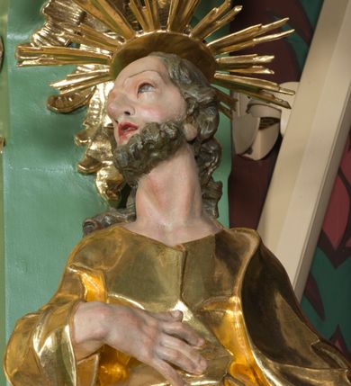 Zdjęcie nr 5: Rzeźba pełna, na cokole, ukazująca św. Józefa w całej postaci, w kontrapoście, frontalnie. Święty ma głowę zwróconą w prawo, prawą rękę przyciska do ciała, w lewej trzyma kwitnącą laskę. Ma prostokątną twarz o wąskich brwiach, dużych, migdałowatych oczach, dużym nosie i rozchylonych ustach, okoloną krótką brodą i układającymi się w pukle, długimi włosami. Jest ubrany w długą suknię w dużym dekoltem, która okleja ciało, łamiąc się grubymi fałdami. Na nogach ma sandały. Polichromia w partiach ciała naturalistyczna, szaty złocone, laska w kolorze brązowym, rozkwitające z niej kwiaty czerwone, na zielonych gałązkach.