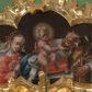 Zdjęcie nr 1: Obraz w kształcie czworoliścia z przedstawieniem Świętej Rodziny. Na środku sceny mały Jezus, widoczny w całej postaci, siedzi na poduszce umieszczonej na postumencie. Patrzy przed siebie, lewą ręką obejmuje św. Józefa, prawą bawi się jego brodą. Jest nagi, między nogami przechodzi mu jedynie biała tkanina. Ma podłużną twarz z wysokim czołem, szeroko rozstawionymi oczami, małym nosem i ustami. Jego jasne włosy kręcą się w loki; wokół głowy nimb. Święty Józef widoczny do pasa, stoi za postumentem. Patrzy na Dzieciątko, prawą ręką przytrzymuje jego plecy, w lewej, uniesionej trzyma kwiat lilii. Jest ubrany w białą, rozciętą pod szyją suknię i żółty płaszcz. Stojąca z lewej strony kompozycji Maria, również widoczna do pasa, spogląda na plecy Dzieciątka. W prawej, uniesionej ręce trzyma kwiat goździka. Ma owalną, młodą twarz, z wysokim czołem, drobnym nosem i ustami. Jest ubrana w czerwoną suknię i niebieski płaszcz. Ma misternie upięte włosy przewiązane czerwoną wstążką, spod niej w lewą stronę rozwiewa się biały welon. Wszystkie tkaniny gną się grubymi fałdami, tło jest jednolicie ciemnobrązowe. 