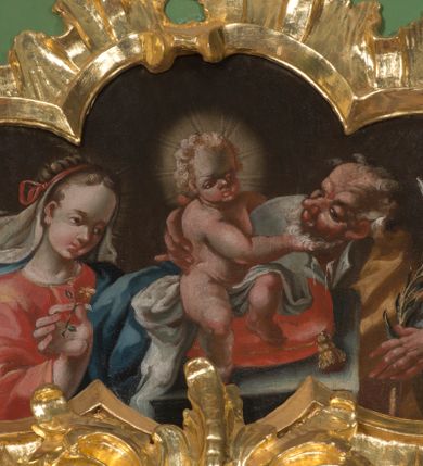 Zdjęcie nr 1: Obraz w kształcie czworoliścia z przedstawieniem Świętej Rodziny. Na środku sceny mały Jezus, widoczny w całej postaci, siedzi na poduszce umieszczonej na postumencie. Patrzy przed siebie, lewą ręką obejmuje św. Józefa, prawą bawi się jego brodą. Jest nagi, między nogami przechodzi mu jedynie biała tkanina. Ma podłużną twarz z wysokim czołem, szeroko rozstawionymi oczami, małym nosem i ustami. Jego jasne włosy kręcą się w loki; wokół głowy nimb. Święty Józef widoczny do pasa, stoi za postumentem. Patrzy na Dzieciątko, prawą ręką przytrzymuje jego plecy, w lewej, uniesionej trzyma kwiat lilii. Jest ubrany w białą, rozciętą pod szyją suknię i żółty płaszcz. Stojąca z lewej strony kompozycji Maria, również widoczna do pasa, spogląda na plecy Dzieciątka. W prawej, uniesionej ręce trzyma kwiat goździka. Ma owalną, młodą twarz, z wysokim czołem, drobnym nosem i ustami. Jest ubrana w czerwoną suknię i niebieski płaszcz. Ma misternie upięte włosy przewiązane czerwoną wstążką, spod niej w lewą stronę rozwiewa się biały welon. Wszystkie tkaniny gną się grubymi fałdami, tło jest jednolicie ciemnobrązowe. 