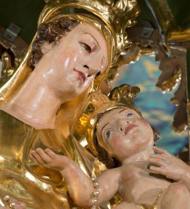 Zdjęcie nr 3: Rzeźba pełna, na cokole, ukazująca Matkę Boską z Dzieciątkiem w całej postaci, w kontrapoście, frontalnie. Maria skłania głowę w kierunku Dzieciątka, które kładzie swoją na jej barku. Maria w prawej ręce trzyma berło, lewą podtrzymuje małego Jezusa, który lewą rękę składa na ciele a prawą wyciąga w kierunku Matki. Maria ma owalną twarz o pełnych policzkach, wąskie brwi, migdałowate, półprzymknięte oczy, duży nos i drobne usta. Jest ubrana w długą suknię i zarzucony na ramiona płaszcz, które łamią się ostrymi fałdami, opadając ku ziemi. Na głowie ma welon, na nogach brązowe buty w szpic. Dzieciątko ma owalną twarz o wąskich brwiach, szeroko rozstawionych, niebieskich oczach, zaróżowionych policzkach i ustach, jest nagie, ma jedynie wąską szarfę owiniętą wokół bioder. Obie postacie mają na głowach otwarte korony utworzone z liści akantu. Polichromia w partiach ciała naturalistyczna, szaty i atrybuty złocone. 