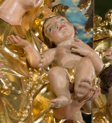 Zdjęcie nr 5: Rzeźba pełna, na cokole, ukazująca Matkę Boską z Dzieciątkiem w całej postaci, w kontrapoście, frontalnie. Maria skłania głowę w kierunku Dzieciątka, które kładzie swoją na jej barku. Maria w prawej ręce trzyma berło, lewą podtrzymuje małego Jezusa, który lewą rękę składa na ciele a prawą wyciąga w kierunku Matki. Maria ma owalną twarz o pełnych policzkach, wąskie brwi, migdałowate, półprzymknięte oczy, duży nos i drobne usta. Jest ubrana w długą suknię i zarzucony na ramiona płaszcz, które łamią się ostrymi fałdami, opadając ku ziemi. Na głowie ma welon, na nogach brązowe buty w szpic. Dzieciątko ma owalną twarz o wąskich brwiach, szeroko rozstawionych, niebieskich oczach, zaróżowionych policzkach i ustach, jest nagie, ma jedynie wąską szarfę owiniętą wokół bioder. Obie postacie mają na głowach otwarte korony utworzone z liści akantu. Polichromia w partiach ciała naturalistyczna, szaty i atrybuty złocone. 