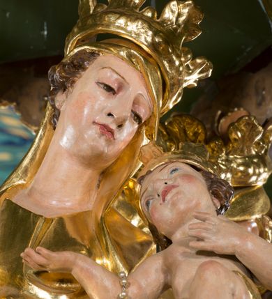Zdjęcie nr 4: Rzeźba pełna, na cokole, ukazująca Matkę Boską z Dzieciątkiem w całej postaci, w kontrapoście, frontalnie. Maria skłania głowę w kierunku Dzieciątka, które kładzie swoją na jej barku. Maria w prawej ręce trzyma berło, lewą podtrzymuje małego Jezusa, który lewą rękę składa na ciele a prawą wyciąga w kierunku Matki. Maria ma owalną twarz o pełnych policzkach, wąskie brwi, migdałowate, półprzymknięte oczy, duży nos i drobne usta. Jest ubrana w długą suknię i zarzucony na ramiona płaszcz, które łamią się ostrymi fałdami, opadając ku ziemi. Na głowie ma welon, na nogach brązowe buty w szpic. Dzieciątko ma owalną twarz o wąskich brwiach, szeroko rozstawionych, niebieskich oczach, zaróżowionych policzkach i ustach, jest nagie, ma jedynie wąską szarfę owiniętą wokół bioder. Obie postacie mają na głowach otwarte korony utworzone z liści akantu. Polichromia w partiach ciała naturalistyczna, szaty i atrybuty złocone. 