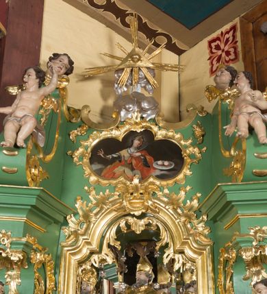 Zdjęcie nr 2: Obraz w kształcie czworoliścia z wizerunkiem św. Agaty ukazanej w półpostaci, frontalnie. Święta spogląda w górę, prawą rękę ma odwiedzioną do boku, w lewej, złożonej na piersi trzyma palmę męczeństwa. Ma pociągłą twarz, z głęboko osadzonymi oczami, długim nosem, mięsistymi ustami i odznaczającym się podbródkiem. W ciemne, długie włosy ma wplecione sznury pereł, na głowie diadem, wokół promieniejący nimb. Jest ubrana w spodnią niebieską suknię z dłuższymi rękawami i wierzchnią, białą, lamowaną złotem. Na to ma nałożony złoty gorset zdobiony perłami i zarzucony czerwony płaszcz, który obszernymi fałdami owija się wokół nóg, pleców oraz przechodzi przez lewą rękę. Po jej lewej stronie, na chmurach, unosi się misa z dwiema obciętymi, krwawiącymi piersiami. Świętą ukazano na tle brunatnego nieba, z szarymi obłokami w dolnej części płótna. 