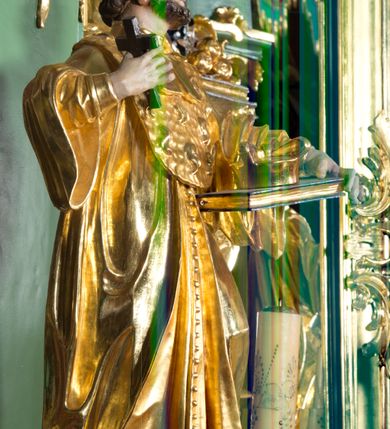 Zdjęcie nr 2: Rzeźba pełna, na cokole ukazująca św. Jana Nepomucena w całej postaci, w kontrapoście z ciałem skręconym lekko w lewą stronę. Święty wznosi wzrok do góry, w prawej, uniesionej ręce trzyma krucyfiks, lewą przytrzymuje opartą na biodrze, zamkniętą księgę. Ma kwadratową twarz o wąskich brwiach, dużych, migdałowatych oczach, dużym nosie i wyraźnie zarysowanych ustach, okoloną krótką brodą i układającymi się w pukle włosami. Jest ubrany w strój kanonika: ma narzuconą na sutannę futrzaną almucję, pod szyją befkę – formę krótkiego krawata. Należący do stroju biret leży na księdze; na nogach ma buty o ściętych czubkach. Polichromia w partiach ciała naturalistyczna, szaty złocone, atrybuty złocone i srebrzone, krzyż brązowy. 