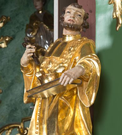 Zdjęcie nr 1: Rzeźba pełna, na cokole ukazująca św. Jana Nepomucena w całej postaci, w kontrapoście z ciałem skręconym lekko w lewą stronę. Święty wznosi wzrok do góry, w prawej, uniesionej ręce trzyma krucyfiks, lewą przytrzymuje opartą na biodrze, zamkniętą księgę. Ma kwadratową twarz o wąskich brwiach, dużych, migdałowatych oczach, dużym nosie i wyraźnie zarysowanych ustach, okoloną krótką brodą i układającymi się w pukle włosami. Jest ubrany w strój kanonika: ma narzuconą na sutannę futrzaną almucję, pod szyją befkę – formę krótkiego krawata. Należący do stroju biret leży na księdze; na nogach ma buty o ściętych czubkach. Polichromia w partiach ciała naturalistyczna, szaty złocone, atrybuty złocone i srebrzone, krzyż brązowy. 