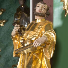 Zdjęcie nr 1: Rzeźba pełna, na cokole ukazująca św. Jana Nepomucena w całej postaci, w kontrapoście z ciałem skręconym lekko w lewą stronę. Święty wznosi wzrok do góry, w prawej, uniesionej ręce trzyma krucyfiks, lewą przytrzymuje opartą na biodrze, zamkniętą księgę. Ma kwadratową twarz o wąskich brwiach, dużych, migdałowatych oczach, dużym nosie i wyraźnie zarysowanych ustach, okoloną krótką brodą i układającymi się w pukle włosami. Jest ubrany w strój kanonika: ma narzuconą na sutannę futrzaną almucję, pod szyją befkę – formę krótkiego krawata. Należący do stroju biret leży na księdze; na nogach ma buty o ściętych czubkach. Polichromia w partiach ciała naturalistyczna, szaty złocone, atrybuty złocone i srebrzone, krzyż brązowy. 