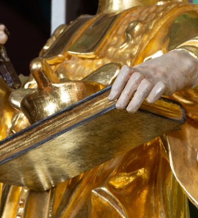 Zdjęcie nr 6: Rzeźba pełna, na cokole ukazująca św. Jana Nepomucena w całej postaci, w kontrapoście z ciałem skręconym lekko w lewą stronę. Święty wznosi wzrok do góry, w prawej, uniesionej ręce trzyma krucyfiks, lewą przytrzymuje opartą na biodrze, zamkniętą księgę. Ma kwadratową twarz o wąskich brwiach, dużych, migdałowatych oczach, dużym nosie i wyraźnie zarysowanych ustach, okoloną krótką brodą i układającymi się w pukle włosami. Jest ubrany w strój kanonika: ma narzuconą na sutannę futrzaną almucję, pod szyją befkę – formę krótkiego krawata. Należący do stroju biret leży na księdze; na nogach ma buty o ściętych czubkach. Polichromia w partiach ciała naturalistyczna, szaty złocone, atrybuty złocone i srebrzone, krzyż brązowy. 