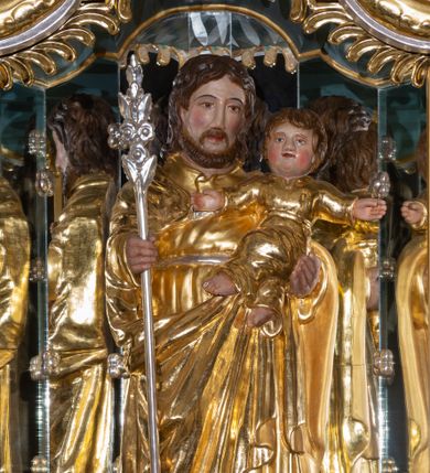 Zdjęcie nr 1: Rzeźba pełna, ustawiona na prostokątnym cokole. Święty Józef ukazany frontalnie, z Dzieciątkiem na lewym ręku, z różdżką Jessego w prawej ręce. Jest ubrany w długą suknię z długimi rękawami, przepasaną sznurkiem oraz długi płaszcz spięty pod szyją, z prawym końcem zarzuconym na lewe przedramię, na stopach sandały. Ma podłużną, okoloną brodą twarz z długim, wąskim nosem i małymi oczami. Włosy brązowe, średniej długości, opadają na plecy. Dzieciątko zostało ukazane frontalnie, z rozpostartymi szeroko rękami, z lewą nogą założoną na prawą. Jest ubrane w długą sukienkę z długimi rękawami, przepasaną w pasie sznurkiem. Ma okrągłą twarz, z rumieńcami na pełnych policzkach i małymi, czerwonymi ustami oraz krótkim, szerokim nosem; włosy krótkie, falowane. Polichromia w partiach ciała naturalistyczna, szaty złocone, atrybut srebrzony.