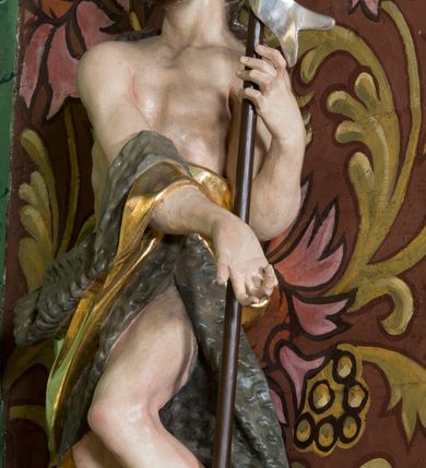 Zdjęcie nr 2: Figura ukazująca św. Jana Chrzciciela w całej postaci, w kontrapoście, sylwetka delikatnie wygięta w lewo, w skręcie. Święty w lewej ręce trzyma długie drzewce zakończone krzyżem z przewiązaną banderolą z napisem „ECCE AGNUS DEI”, prawą rękę silnie wysuwa w lewą stronę. Twarz szczupła o dużych szeroko rozstawionych i migdałowatych oczach z wyraźnie rzeźbionymi powiekami, dużym zaoblonym nosem; okolona zarostem oraz włosami do ramion w postaci silnie przylegających do głowy, falujących pukli. Święty ma narzucony płaszcz ze skóry ze złoconym podbiciem, zawiązany na prawym przedramieniu i w partii bioder, odsłaniający lewe udo. U stóp figury rzeźba leżącego baranka z głową skierowaną ku górze. Polichromia partii ciała naturalistyczna, detal złocony i srebrzony. 