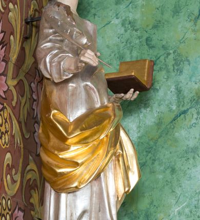 Zdjęcie nr 2: Rzeźba z przedstawieniem św. Jana Ewangelisty. Święty został ukazany w całej postaci frontalnie, w kontrapoście, z sylwetką delikatnie esowato wygiętą, z głową skierowaną w prawo, lekko uniesioną. Obie ręce ma ugięte, rozłożone na boki, w lewej otwarta księga, w prawej pióro. Twarz szczupła, o młodzieńczych rysach, oczach uniesionych ku górze, okolona długimi włosami w postaci głęboko rzeźbionych, wijących się pukli. Ubrany jest w długą tunikę, silnie przylegającą do ciała oraz płaszcz przewiązany przez lewe ramię i zawinięty na prawym boku i udzie. Przy lewej nodze figurka orła. Polichromia naturalistyczna w odsłoniętych partiach ciała, szaty i atrybuty złocone i srebrzone.