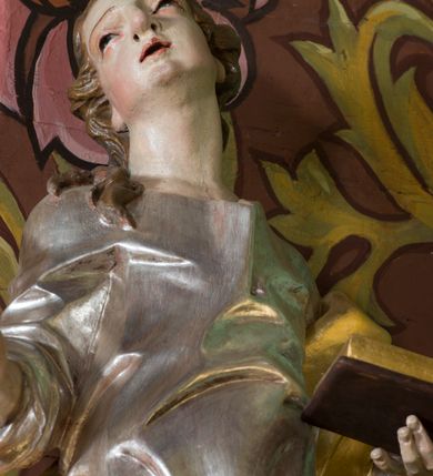 Zdjęcie nr 6: Rzeźba z przedstawieniem św. Jana Ewangelisty. Święty został ukazany w całej postaci frontalnie, w kontrapoście, z sylwetką delikatnie esowato wygiętą, z głową skierowaną w prawo, lekko uniesioną. Obie ręce ma ugięte, rozłożone na boki, w lewej otwarta księga, w prawej pióro. Twarz szczupła, o młodzieńczych rysach, oczach uniesionych ku górze, okolona długimi włosami w postaci głęboko rzeźbionych, wijących się pukli. Ubrany jest w długą tunikę, silnie przylegającą do ciała oraz płaszcz przewiązany przez lewe ramię i zawinięty na prawym boku i udzie. Przy lewej nodze figurka orła. Polichromia naturalistyczna w odsłoniętych partiach ciała, szaty i atrybuty złocone i srebrzone.