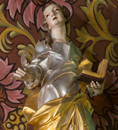 Zdjęcie nr 1: Rzeźba z przedstawieniem św. Jana Ewangelisty. Święty został ukazany w całej postaci frontalnie, w kontrapoście, z sylwetką delikatnie esowato wygiętą, z głową skierowaną w prawo, lekko uniesioną. Obie ręce ma ugięte, rozłożone na boki, w lewej otwarta księga, w prawej pióro. Twarz szczupła, o młodzieńczych rysach, oczach uniesionych ku górze, okolona długimi włosami w postaci głęboko rzeźbionych, wijących się pukli. Ubrany jest w długą tunikę, silnie przylegającą do ciała oraz płaszcz przewiązany przez lewe ramię i zawinięty na prawym boku i udzie. Przy lewej nodze figurka orła. Polichromia naturalistyczna w odsłoniętych partiach ciała, szaty i atrybuty złocone i srebrzone.