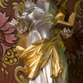 Zdjęcie nr 1: Rzeźba z przedstawieniem św. Jana Ewangelisty. Święty został ukazany w całej postaci frontalnie, w kontrapoście, z sylwetką delikatnie esowato wygiętą, z głową skierowaną w prawo, lekko uniesioną. Obie ręce ma ugięte, rozłożone na boki, w lewej otwarta księga, w prawej pióro. Twarz szczupła, o młodzieńczych rysach, oczach uniesionych ku górze, okolona długimi włosami w postaci głęboko rzeźbionych, wijących się pukli. Ubrany jest w długą tunikę, silnie przylegającą do ciała oraz płaszcz przewiązany przez lewe ramię i zawinięty na prawym boku i udzie. Przy lewej nodze figurka orła. Polichromia naturalistyczna w odsłoniętych partiach ciała, szaty i atrybuty złocone i srebrzone.