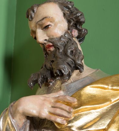 Zdjęcie nr 4: Rzeźba przedstawiająca św. Pawła w całej postaci. Figura ustawiona frontalnie, w kontrapoście, z głową pochyloną do przodu, prawą ręką spoczywającą na piersi, lewą wyciągniętą w bok, trzymającą miecz zwrócony ostrzem w dół. Twarz szczupła z wysokim czołem, o wyraźnie widocznych łukach brwiowych, dużym, zaoblonym nosie i zaznaczonych kościach policzkowych, okolona długą brodą silnie skręconą w pukle i głęboko rzeźbioną oraz krótkimi, silnie skręconymi włosami. Święty jest ubrany w długą suknię przewiązaną w talii oraz płaszcz podtrzymywany na piersi, przerzucony przez lewe ramię. Polichromia naturalistyczna w odsłoniętych partiach ciała, szaty i atrybuty złocone i srebrzone.