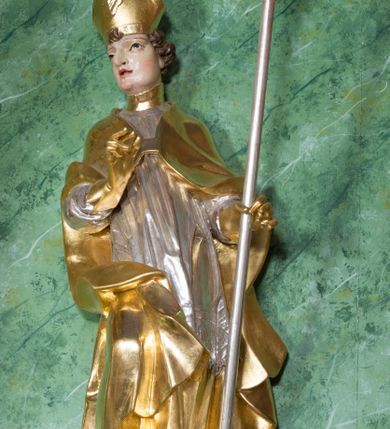 Zdjęcie nr 2: Rzeźba ukazująca św. Stanisława w całej postaci. Figura ustawiona frontalnie, w kontrapoście, łukowato przechylona w lewo; w lewej ręce trzyma pastorał, prawa ugięta, wyciągnięta przed siebie. Twarz kwadratowa o silnie zaznaczonych liniach łuków brwiowych i głęboko rzeźbionych powiekach, dużych oczach i nosie oraz małych, rozchylonych ustach, okolona krótkimi, silnie skręconymi włosami. Święty ubrany jest w złoconą albę, srebrzoną rokietę oraz złoconą kapę zapiętą na piersi, opadającą na ramiona, z fragmentem tkaniny zarzuconym na lewą nogę. Polichromia naturalistyczna w odsłoniętych partiach ciała, szaty i atrybuty złocone i srebrzone.