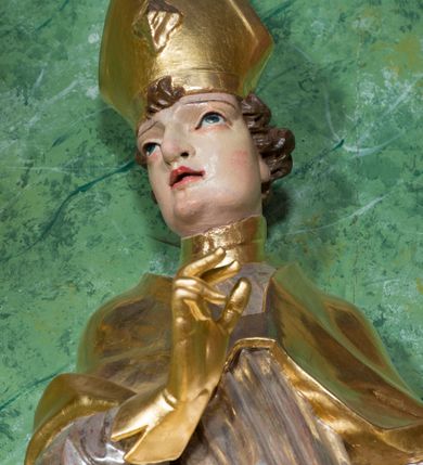 Zdjęcie nr 5: Rzeźba ukazująca św. Stanisława w całej postaci. Figura ustawiona frontalnie, w kontrapoście, łukowato przechylona w lewo; w lewej ręce trzyma pastorał, prawa ugięta, wyciągnięta przed siebie. Twarz kwadratowa o silnie zaznaczonych liniach łuków brwiowych i głęboko rzeźbionych powiekach, dużych oczach i nosie oraz małych, rozchylonych ustach, okolona krótkimi, silnie skręconymi włosami. Święty ubrany jest w złoconą albę, srebrzoną rokietę oraz złoconą kapę zapiętą na piersi, opadającą na ramiona, z fragmentem tkaniny zarzuconym na lewą nogę. Polichromia naturalistyczna w odsłoniętych partiach ciała, szaty i atrybuty złocone i srebrzone.