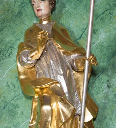 Zdjęcie nr 3: Rzeźba ukazująca św. Stanisława w całej postaci. Figura ustawiona frontalnie, w kontrapoście, łukowato przechylona w lewo; w lewej ręce trzyma pastorał, prawa ugięta, wyciągnięta przed siebie. Twarz kwadratowa o silnie zaznaczonych liniach łuków brwiowych i głęboko rzeźbionych powiekach, dużych oczach i nosie oraz małych, rozchylonych ustach, okolona krótkimi, silnie skręconymi włosami. Święty ubrany jest w złoconą albę, srebrzoną rokietę oraz złoconą kapę zapiętą na piersi, opadającą na ramiona, z fragmentem tkaniny zarzuconym na lewą nogę. Polichromia naturalistyczna w odsłoniętych partiach ciała, szaty i atrybuty złocone i srebrzone.