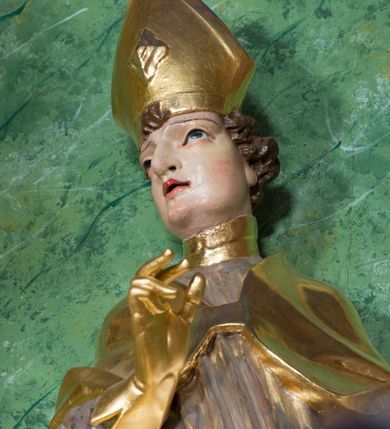 Zdjęcie nr 6: Rzeźba ukazująca św. Stanisława w całej postaci. Figura ustawiona frontalnie, w kontrapoście, łukowato przechylona w lewo; w lewej ręce trzyma pastorał, prawa ugięta, wyciągnięta przed siebie. Twarz kwadratowa o silnie zaznaczonych liniach łuków brwiowych i głęboko rzeźbionych powiekach, dużych oczach i nosie oraz małych, rozchylonych ustach, okolona krótkimi, silnie skręconymi włosami. Święty ubrany jest w złoconą albę, srebrzoną rokietę oraz złoconą kapę zapiętą na piersi, opadającą na ramiona, z fragmentem tkaniny zarzuconym na lewą nogę. Polichromia naturalistyczna w odsłoniętych partiach ciała, szaty i atrybuty złocone i srebrzone.