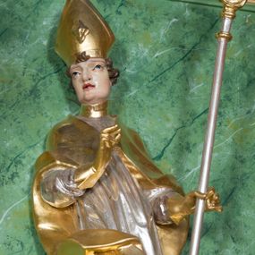 Zdjęcie nr 1: Rzeźba ukazująca św. Stanisława w całej postaci. Figura ustawiona frontalnie, w kontrapoście, łukowato przechylona w lewo; w lewej ręce trzyma pastorał, prawa ugięta, wyciągnięta przed siebie. Twarz kwadratowa o silnie zaznaczonych liniach łuków brwiowych i głęboko rzeźbionych powiekach, dużych oczach i nosie oraz małych, rozchylonych ustach, okolona krótkimi, silnie skręconymi włosami. Święty ubrany jest w złoconą albę, srebrzoną rokietę oraz złoconą kapę zapiętą na piersi, opadającą na ramiona, z fragmentem tkaniny zarzuconym na lewą nogę. Polichromia naturalistyczna w odsłoniętych partiach ciała, szaty i atrybuty złocone i srebrzone.