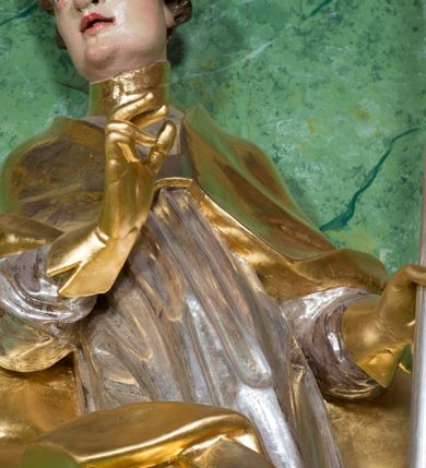 Zdjęcie nr 4: Rzeźba ukazująca św. Stanisława w całej postaci. Figura ustawiona frontalnie, w kontrapoście, łukowato przechylona w lewo; w lewej ręce trzyma pastorał, prawa ugięta, wyciągnięta przed siebie. Twarz kwadratowa o silnie zaznaczonych liniach łuków brwiowych i głęboko rzeźbionych powiekach, dużych oczach i nosie oraz małych, rozchylonych ustach, okolona krótkimi, silnie skręconymi włosami. Święty ubrany jest w złoconą albę, srebrzoną rokietę oraz złoconą kapę zapiętą na piersi, opadającą na ramiona, z fragmentem tkaniny zarzuconym na lewą nogę. Polichromia naturalistyczna w odsłoniętych partiach ciała, szaty i atrybuty złocone i srebrzone.