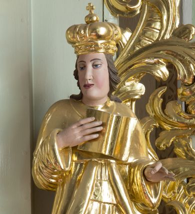 Zdjęcie nr 1: Rzeźba przedstawia nierozpoznanego świętego królewicza węgierskiego, ustawionego na półkolistym, srebrzonym cokole. Święty ukazany frontalnie, w prawej dłoni trzyma otwartą księgę, a lewą, ugiętą w łokciu wyciąga do trzymania niezachowanego atrybutu. Twarz pełna, pulchna w dolnej części o delikatnie rozchylonych ustach i dużych oczach. Głowa okolona długimi, ciemnobrązowymi włosami, zawiniętymi w duże loki do tyłu. Święty ubrany jest w długą suknię i płaszcz, na głowie ma założoną mitrę książęcą. Dłonie wyrzeźbione sztywno i niedbale. Polichromia w odsłoniętych partiach ciała naturalistyczna, szaty i atrybuty złocone, cokół srebrzony.




