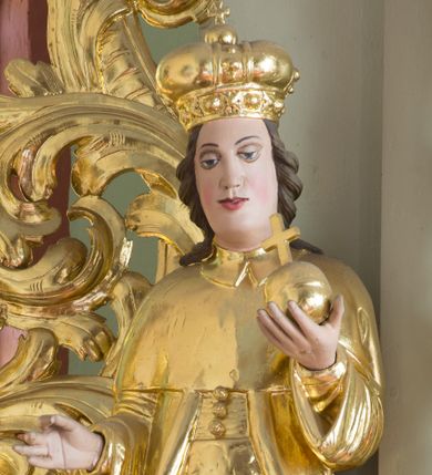 Zdjęcie nr 1: Rzeźba przedstawia nierozpoznanego świętego królewicza węgierskiego, ustawionego na półkolistym, srebrzonym cokole. Święty ukazany frontalnie, w lewej dłoni trzyma jabłko królewskie, a prawą, ugiętą w łokciu wyciąga do trzymania niezachowanego atrybutu. Twarz pełna, pulchna w dolnej części, o delikatnie rozchylonych ustach i dużych oczach. Głowa okolona długimi, ciemnobrązowymi włosami, zawiniętymi w duże loki do tyłu. Święty ubrany w spodnie, wysokie buty z cholewami, tunikę, pas i płaszcz, na głowie ma założoną mitrę książęcą. Draperia szat zaznaczona tylko przy rękawach; dłonie wyrzeźbione sztywno i niedbale. Polichromia w odsłoniętych partiach ciała naturalistyczna, szaty i atrybuty złocone oraz srebrzone.


