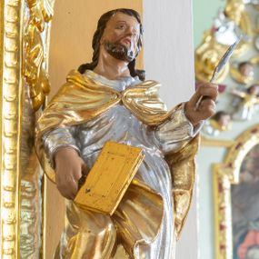 Zdjęcie nr 1: Rzeźba przedstawia świętego ewangelistę ustawionego na półkolistym, brązowym cokole. Święty ukazany frontalnie o delikatnie esowato wygiętej sylwetce, w prawej dłoni trzyma księgę, a w lewej pióro. Twarz o idealizowanych rysach, nos długi i prosty, wzrok wzniesiony ku górze. Głowa okolona krótką i brązową brodą oraz długimi włosami. Święty ubrany jest w srebrzoną suknię oraz złocony płaszcz, spięty na piersi, na stopach ma założone złocone buty. Draperia szat bujna o silnie falującym brzegu płaszcza, dłonie wyrzeźbione sztywno i niedbale. Polichromia w odsłoniętych partiach ciała naturalistyczna, szaty i atrybuty złocone oraz srebrzone.