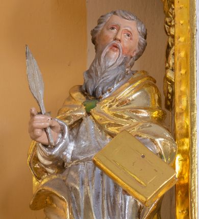 Zdjęcie nr 1: Rzeźba przedstawia świętego ewangelistę ustawionego na półkolistym, brązowym cokole. Święty ukazany frontalnie o delikatnie esowato wygiętej sylwetce. W prawej dłoni trzyma pióro, a w lewej księgę. Twarz o idealizowanych rysach, nos długi i prosty, wzrok wzniesiony ku górze. Głowa okolona długą i siwą brodą oraz siwymi, średniej długości włosami. Święty ubrany jest w srebrzoną suknię oraz złocony płaszcz, spięty na piersi zieloną klamrą, na stopach ma założone złocone buty. Draperia szat bujna o silnie falującym brzegu płaszcza. Polichromia w odsłoniętych partiach ciała naturalistyczna, szaty i atrybuty złocone oraz srebrzone.