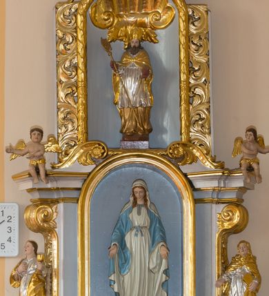 Zdjęcie nr 3: Ołtarz architektoniczny, dwukondygnacyjny, jednoosiowy ze zwieńczeniem. Ołtarz prostopadłościenny z drewnianą mensą i kamiennym portatylem. Antependium ozdobione trzema płycinami z czego środkowa w kształcie czwórliścia przenikającego się z kwadratem; dwie boczne, ustawione antytetycznie, w kształcie leżącego prostokąta z parą boków wewnętrznych w kształcie łuku nadwieszonego wklęsłego. W płycinie środkowej polichromowany monogram maryjny, w płycinach bocznych po lewej stronie Zwiastowanie, a po prawej Ucieczka do Egiptu. 

Nastawa na niewysokim, uskokowym cokole o ukośnie ustawionych bokach, zdobionych słonecznikami. W środkowej partii cokołu wkomponowane tabernakulum w formie prostopadłościennej szafki na profilowanym, uskokowym cokole z narożami ujętymi dwoma parami pilastrów z półwałkiem pośrodku trzonu, wspierających profilowany, uskokowy gzyms. Drzwiczki tabernakulum w kształcie prostokąta zamkniętego łukiem półkolistym, w polu płaskorzeźbiony kielich z Hostią oraz kłosami zbóż. Kondygnacja flankowana parą zdwojonych pilastrów oraz dostawionych, ustawionych pod kątem pilastrów z nałożonymi spływami wolutowymi. Podpory wspierają przerywane, niepełne belkowanie oraz przyczółek. Pole środkowe zajmuje wnęka w kształcie stojącego prostokąta, zamkniętego półkoliście. W polu figura Matki Boskiej Niepokalanie Poczętej. Pod wnęką podwieszona, rzeźbiona girlanda. Po lewej stronie kondygnacji, na cokole ułożonym na zakończeniu woluty ustawiona jest rzeźba św. Jana Ewangelisty, a po drugiej stronie figura nierozpoznanej świętej. Na belkowaniu usadzona jest para aniołków. Druga kondygnacja w formie wnęki w kształcie stojącego prostokąta, zamkniętego półkoliście, flankowana po bokach filarami, ozdobionymi liśćmi akantu. W polu umieszczona rzeźba św. Mikołaja. Nad rzeźbą stylizowana duża muszla palmetowa z podwieszonymi liśćmi akantu. Zwieńczenie w formie promienistej glorii z Okiem Opatrzności pośrodku, ujętej wieńcem srebrzonych obłoków. Struktura polichromowana na biało, detal architektoniczny i ornamentyka złocone oraz srebrzone. Na cokole tabliczka z napisem: „FUNDACJA / SREBRZENIE ŚWIECZNIKÓW / OŁTARZA NMP / Franciszek i Małgorzata Łojek / 2003”. 