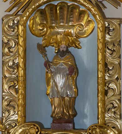 Zdjęcie nr 6: Ołtarz architektoniczny, dwukondygnacyjny, jednoosiowy ze zwieńczeniem. Ołtarz prostopadłościenny z drewnianą mensą i kamiennym portatylem. Antependium ozdobione trzema płycinami z czego środkowa w kształcie czwórliścia przenikającego się z kwadratem; dwie boczne, ustawione antytetycznie, w kształcie leżącego prostokąta z parą boków wewnętrznych w kształcie łuku nadwieszonego wklęsłego. W płycinie środkowej polichromowany monogram maryjny, w płycinach bocznych po lewej stronie Zwiastowanie, a po prawej Ucieczka do Egiptu. 

Nastawa na niewysokim, uskokowym cokole o ukośnie ustawionych bokach, zdobionych słonecznikami. W środkowej partii cokołu wkomponowane tabernakulum w formie prostopadłościennej szafki na profilowanym, uskokowym cokole z narożami ujętymi dwoma parami pilastrów z półwałkiem pośrodku trzonu, wspierających profilowany, uskokowy gzyms. Drzwiczki tabernakulum w kształcie prostokąta zamkniętego łukiem półkolistym, w polu płaskorzeźbiony kielich z Hostią oraz kłosami zbóż. Kondygnacja flankowana parą zdwojonych pilastrów oraz dostawionych, ustawionych pod kątem pilastrów z nałożonymi spływami wolutowymi. Podpory wspierają przerywane, niepełne belkowanie oraz przyczółek. Pole środkowe zajmuje wnęka w kształcie stojącego prostokąta, zamkniętego półkoliście. W polu figura Matki Boskiej Niepokalanie Poczętej. Pod wnęką podwieszona, rzeźbiona girlanda. Po lewej stronie kondygnacji, na cokole ułożonym na zakończeniu woluty ustawiona jest rzeźba św. Jana Ewangelisty, a po drugiej stronie figura nierozpoznanej świętej. Na belkowaniu usadzona jest para aniołków. Druga kondygnacja w formie wnęki w kształcie stojącego prostokąta, zamkniętego półkoliście, flankowana po bokach filarami, ozdobionymi liśćmi akantu. W polu umieszczona rzeźba św. Mikołaja. Nad rzeźbą stylizowana duża muszla palmetowa z podwieszonymi liśćmi akantu. Zwieńczenie w formie promienistej glorii z Okiem Opatrzności pośrodku, ujętej wieńcem srebrzonych obłoków. Struktura polichromowana na biało, detal architektoniczny i ornamentyka złocone oraz srebrzone. Na cokole tabliczka z napisem: „FUNDACJA / SREBRZENIE ŚWIECZNIKÓW / OŁTARZA NMP / Franciszek i Małgorzata Łojek / 2003”. 