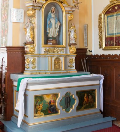 Zdjęcie nr 1: Ołtarz architektoniczny, dwukondygnacyjny, jednoosiowy ze zwieńczeniem. Ołtarz prostopadłościenny z drewnianą mensą i kamiennym portatylem. Antependium ozdobione trzema płycinami z czego środkowa w kształcie czwórliścia przenikającego się z kwadratem; dwie boczne, ustawione antytetycznie, w kształcie leżącego prostokąta z parą boków wewnętrznych w kształcie łuku nadwieszonego wklęsłego. W płycinie środkowej polichromowany monogram maryjny, w płycinach bocznych po lewej stronie Zwiastowanie, a po prawej Ucieczka do Egiptu. 

Nastawa na niewysokim, uskokowym cokole o ukośnie ustawionych bokach, zdobionych słonecznikami. W środkowej partii cokołu wkomponowane tabernakulum w formie prostopadłościennej szafki na profilowanym, uskokowym cokole z narożami ujętymi dwoma parami pilastrów z półwałkiem pośrodku trzonu, wspierających profilowany, uskokowy gzyms. Drzwiczki tabernakulum w kształcie prostokąta zamkniętego łukiem półkolistym, w polu płaskorzeźbiony kielich z Hostią oraz kłosami zbóż. Kondygnacja flankowana parą zdwojonych pilastrów oraz dostawionych, ustawionych pod kątem pilastrów z nałożonymi spływami wolutowymi. Podpory wspierają przerywane, niepełne belkowanie oraz przyczółek. Pole środkowe zajmuje wnęka w kształcie stojącego prostokąta, zamkniętego półkoliście. W polu figura Matki Boskiej Niepokalanie Poczętej. Pod wnęką podwieszona, rzeźbiona girlanda. Po lewej stronie kondygnacji, na cokole ułożonym na zakończeniu woluty ustawiona jest rzeźba św. Jana Ewangelisty, a po drugiej stronie figura nierozpoznanej świętej. Na belkowaniu usadzona jest para aniołków. Druga kondygnacja w formie wnęki w kształcie stojącego prostokąta, zamkniętego półkoliście, flankowana po bokach filarami, ozdobionymi liśćmi akantu. W polu umieszczona rzeźba św. Mikołaja. Nad rzeźbą stylizowana duża muszla palmetowa z podwieszonymi liśćmi akantu. Zwieńczenie w formie promienistej glorii z Okiem Opatrzności pośrodku, ujętej wieńcem srebrzonych obłoków. Struktura polichromowana na biało, detal architektoniczny i ornamentyka złocone oraz srebrzone. Na cokole tabliczka z napisem: „FUNDACJA / SREBRZENIE ŚWIECZNIKÓW / OŁTARZA NMP / Franciszek i Małgorzata Łojek / 2003”. 
